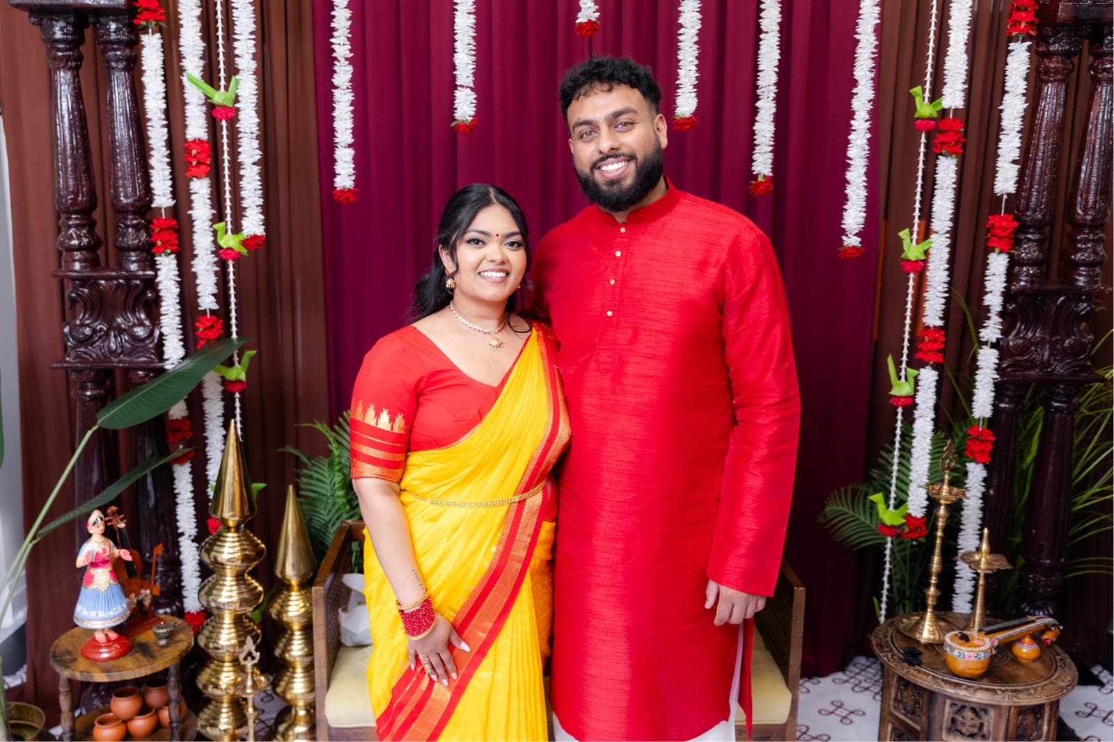 Piratheepan and Shivanna smiling together in matching red and yellow traditional outfits in front of a floral ceremony backdrop