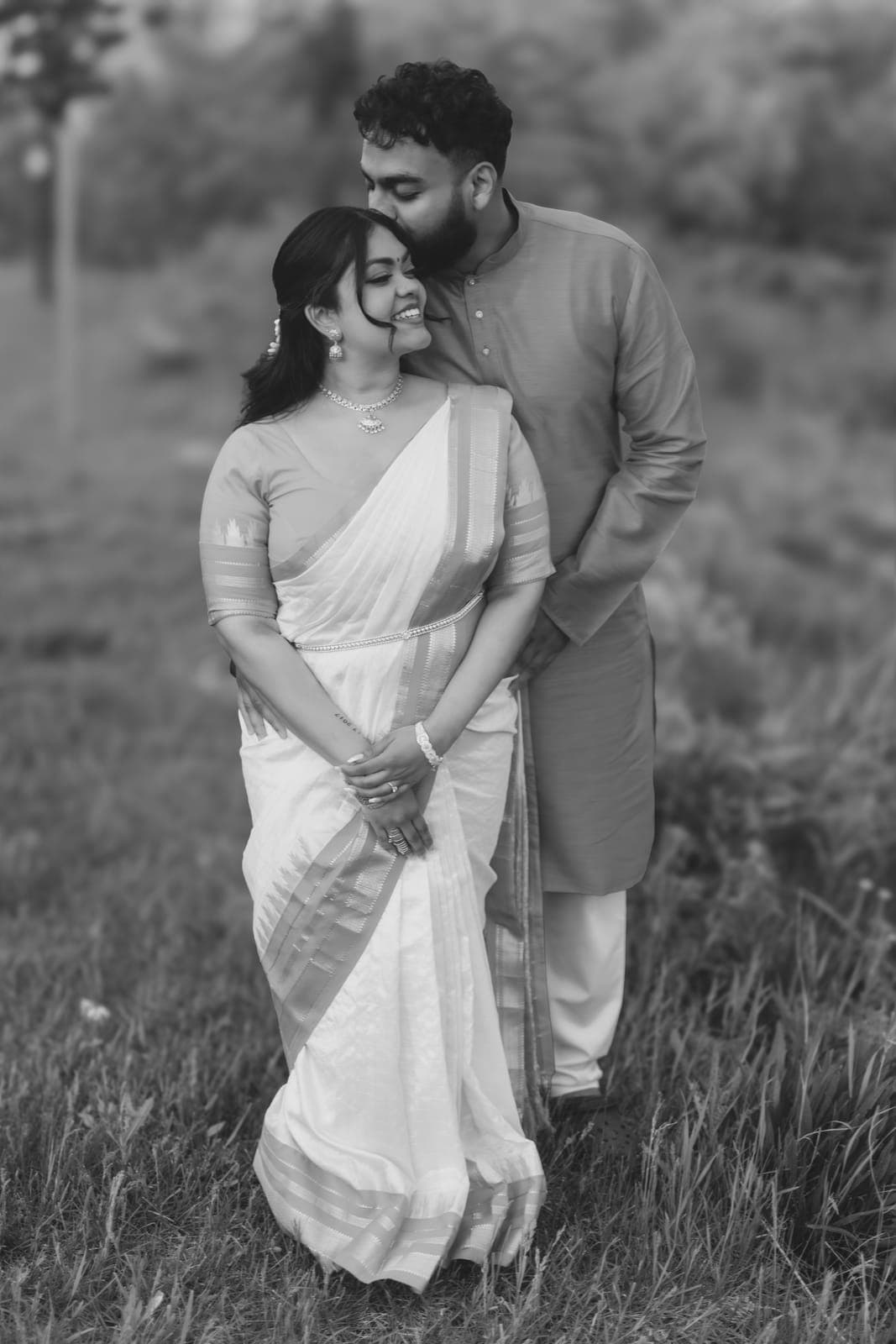 Black-and-white portrait of Piratheepan kissing Shivanna's forehead in a white and gold saree, standing in a grassy field