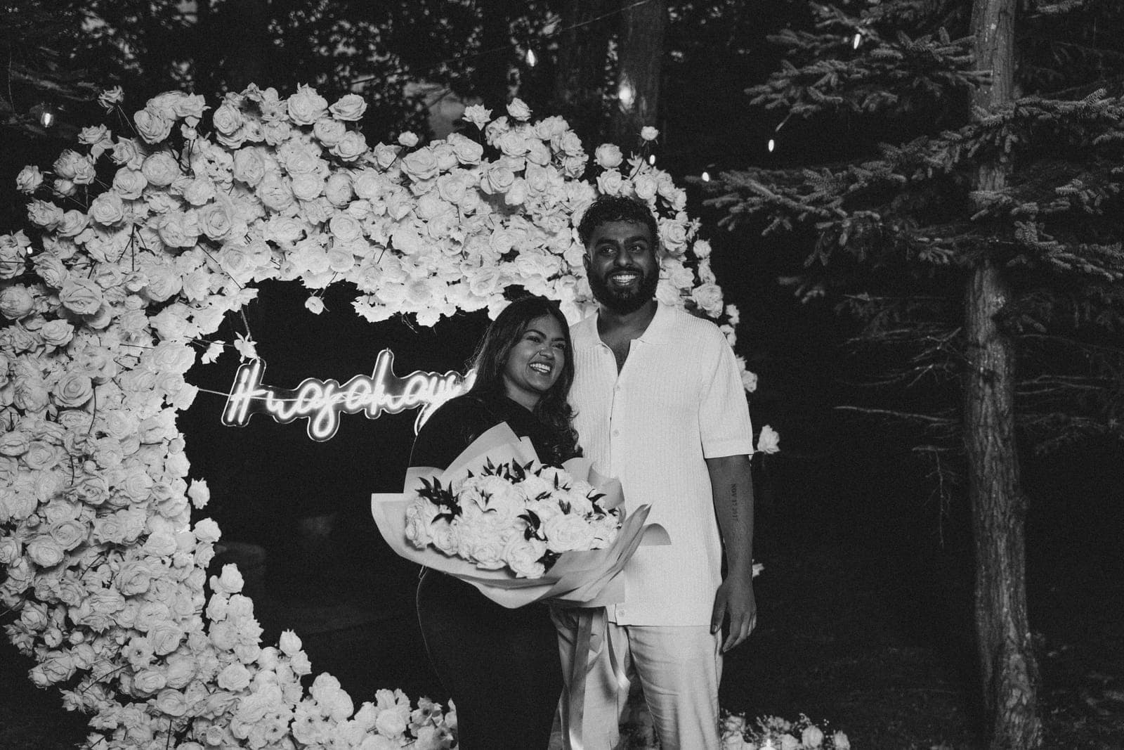 Black-and-white shot of Piratheepan and Shivanna laughing together under the rose-covered proposal arch, neon sign glowing behind them